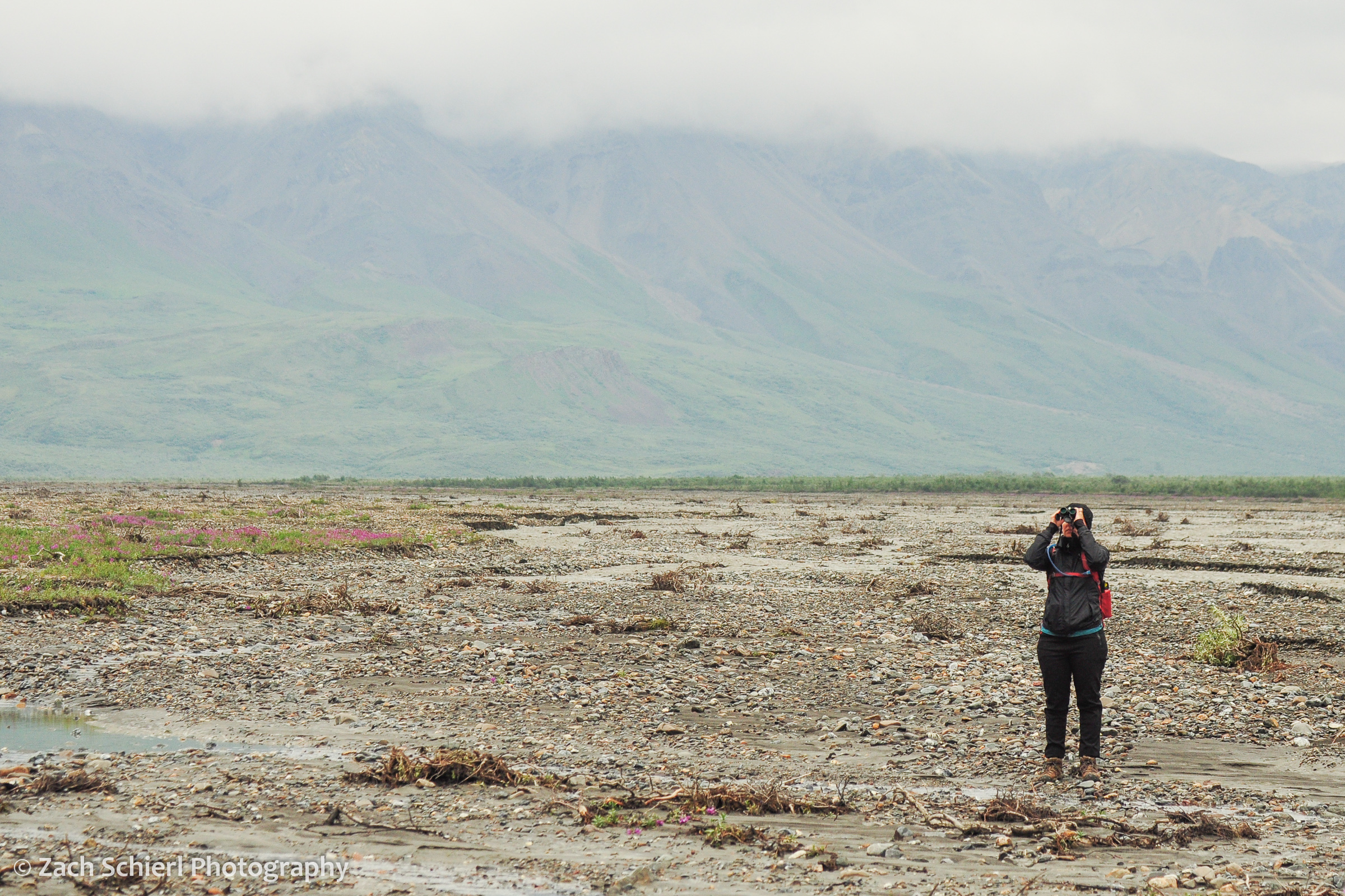 A tourist stand alongside a river scanning the mountains with binoculars