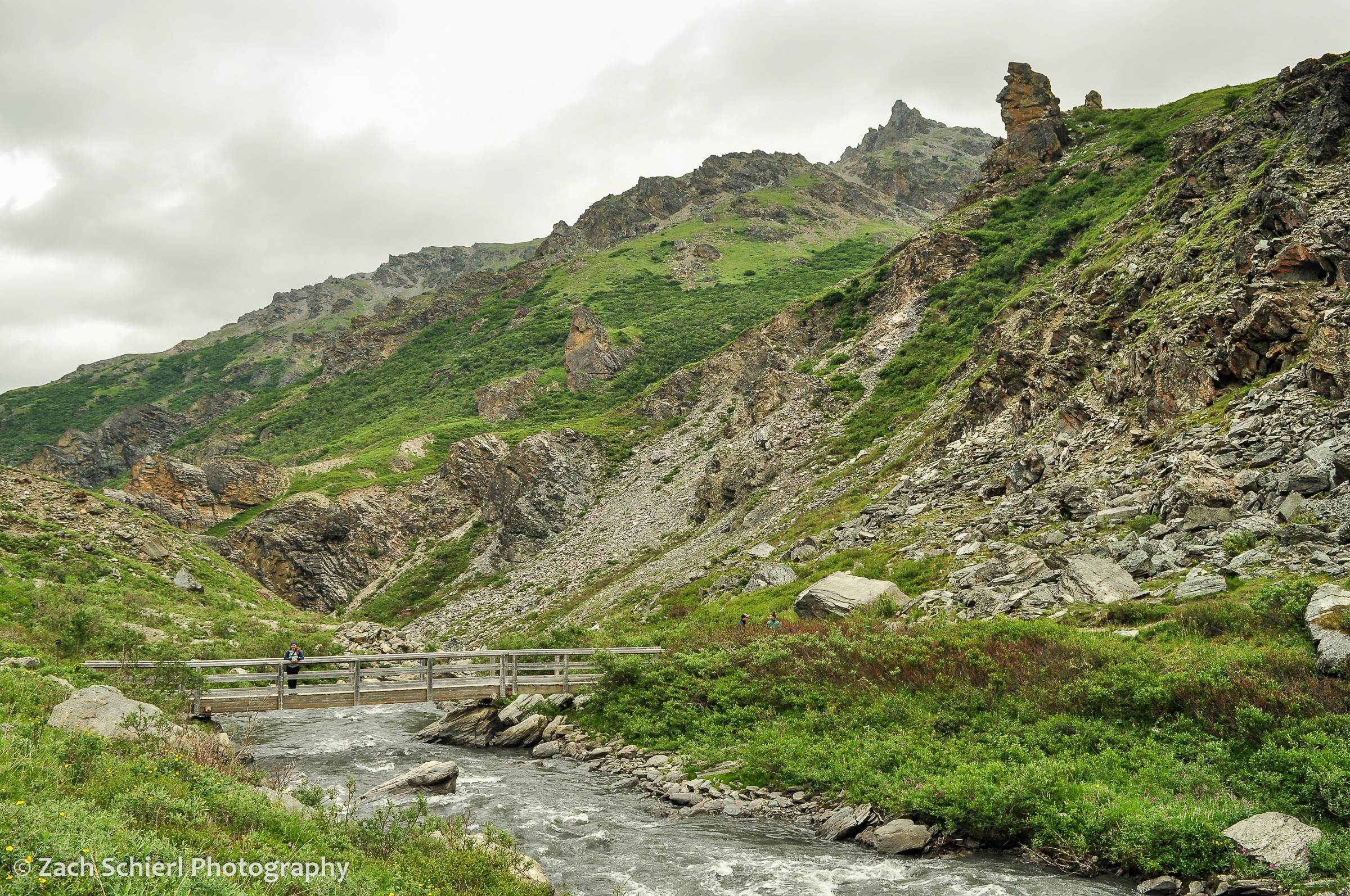 A river flows through rocky crags, as someone stands on a bridge over the river