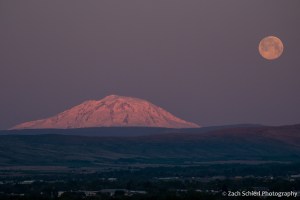 Pink morning light on a snow-capped mountain peak with a full moon setting in the background. 