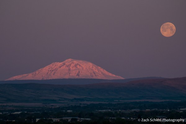 Pink morning light on a snow-capped mountain peak with a full moon setting in the background.