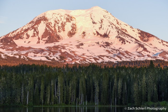 Orange and pink sunset light on the summit of a tall snow-capped mountain.
