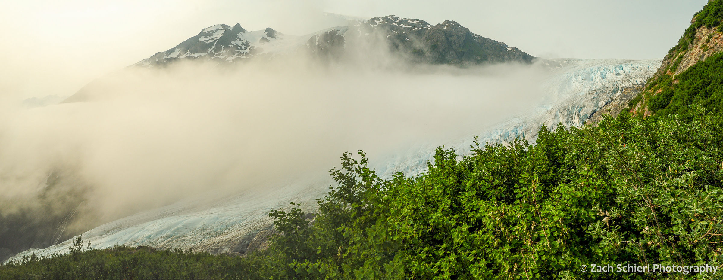A valley containing a glacier is partially obscure by a bank of clouds