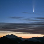 A comet and its tail appears in the pre-dawn sky with a mountain range and valley fog in the foreground
