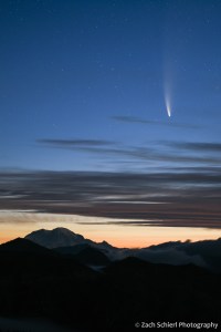 A comet and its tail appears in the pre-dawn sky with a mountain range and valley fog in the foreground