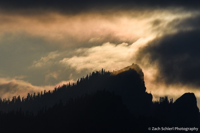 The setting sun casts a pink glow on low clouds above a rocky pinnacle with a lookout tower. 