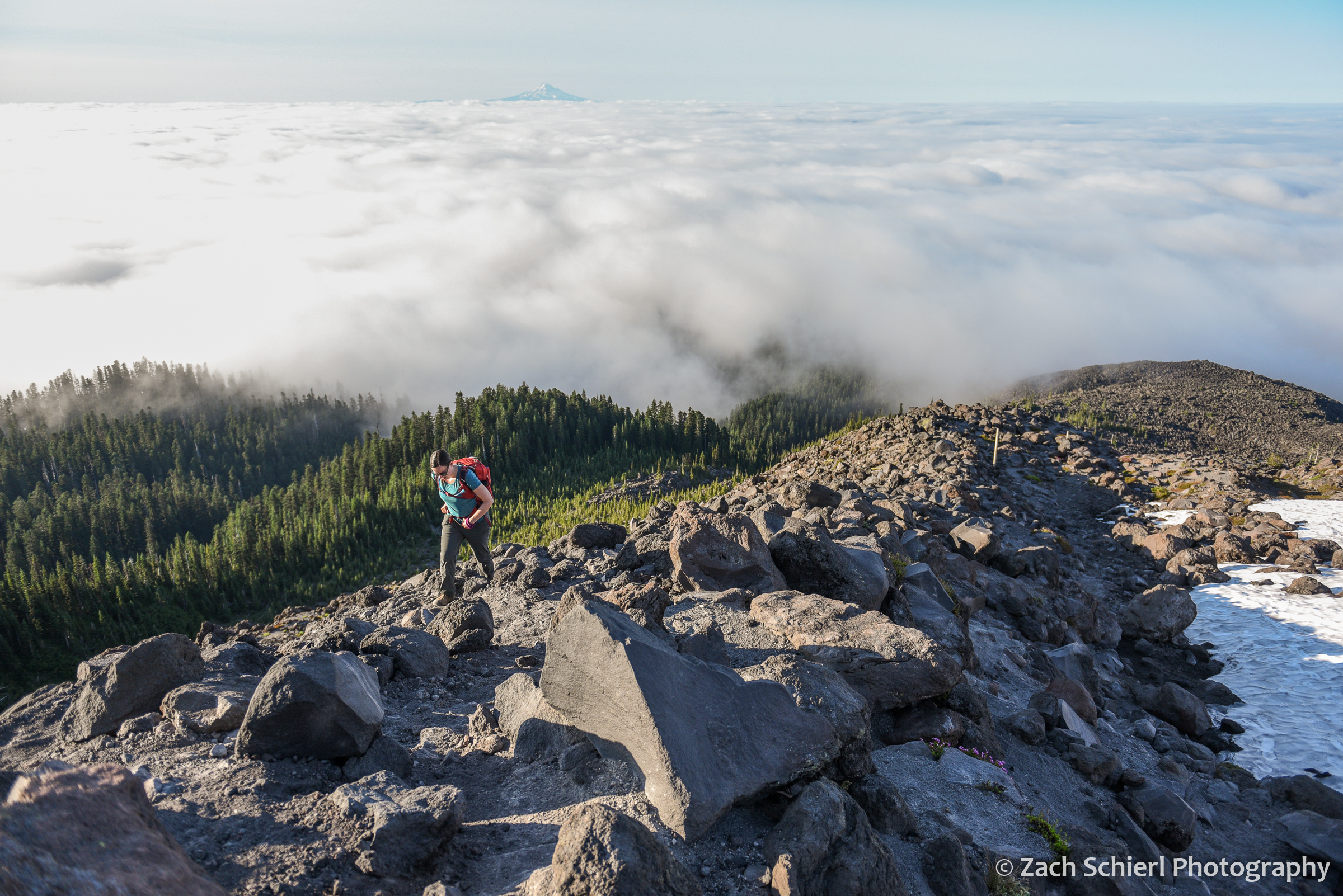 A hiker walks along a rocky ridge with clouds and a distant volcanic peak in the backgrounde background