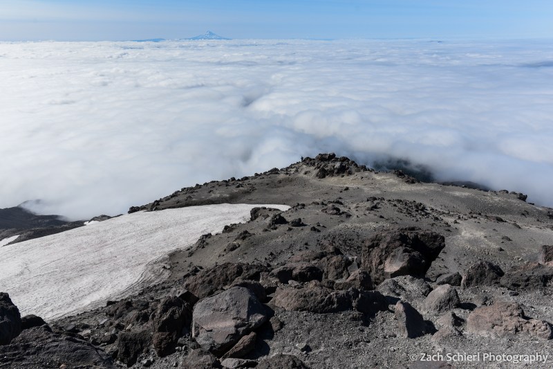 Dark, jagged volcanic rocks on the slopes of Mt. St. Helens with clouds far below
