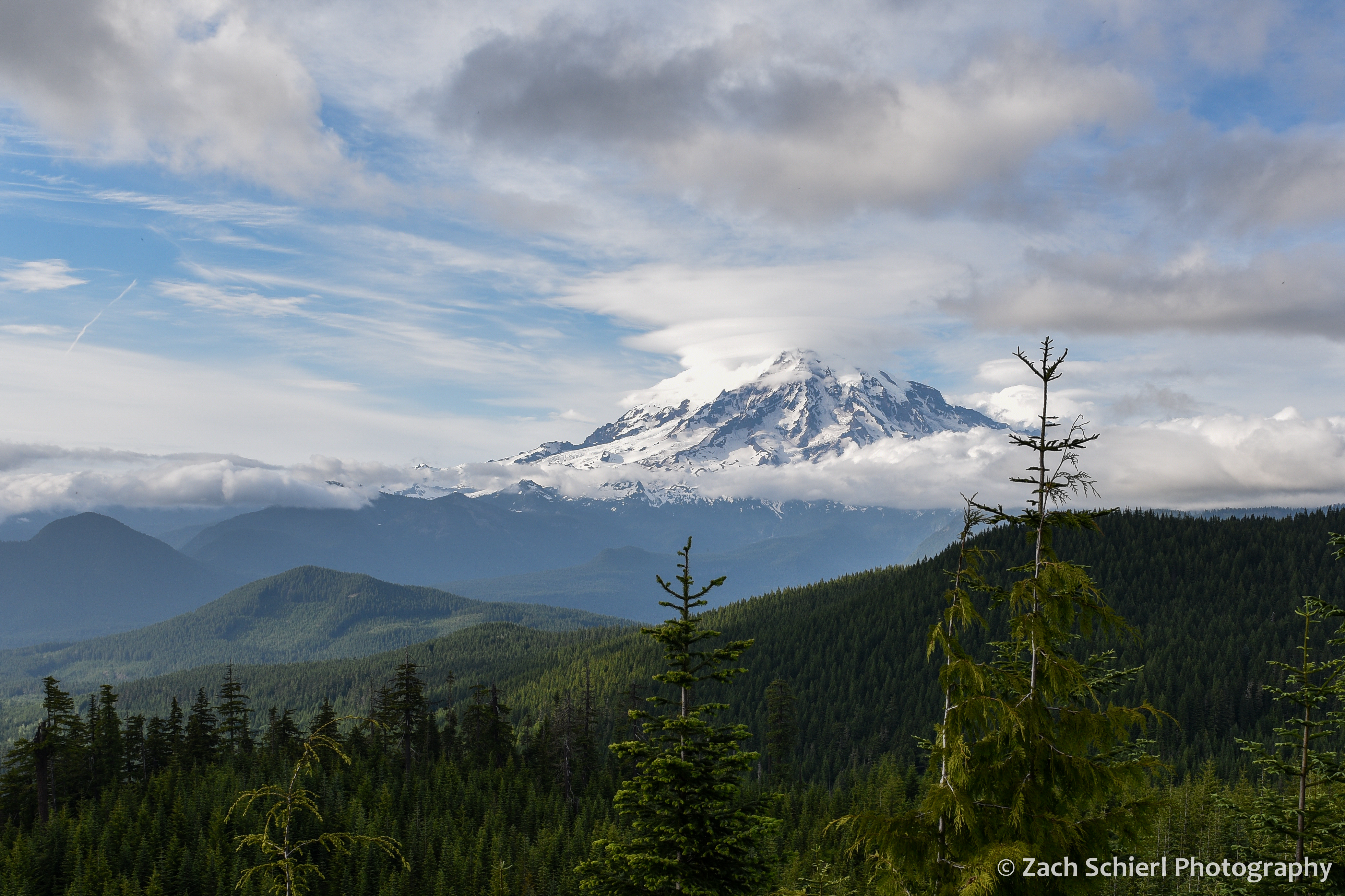 A green forested valley with a conical volcanic peak in the background. Many different types of clouds in the sky.