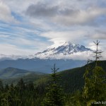 A green forested valley with a conical volcanic peak in the background. Many different types of clouds in the sky.