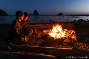Two people sit on a log illuminated by the glow of a campfire. 