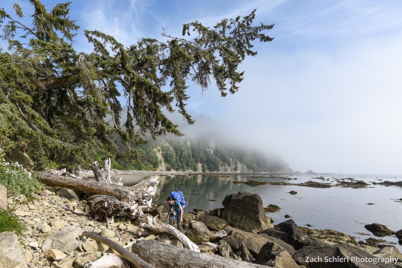 A hiker with a large backpack navigates a pile of boulders along a coastline.