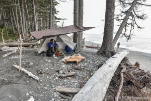 A camper sits next to a tent under a tarp, scanning the skies with binoculars.