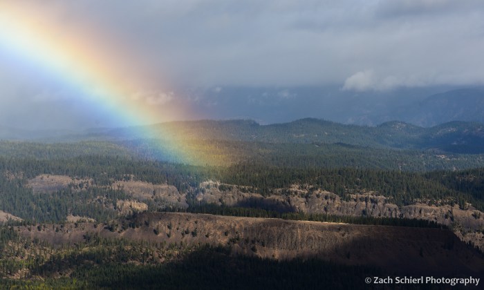 A bright rainbow intersects a series of forested plateaus