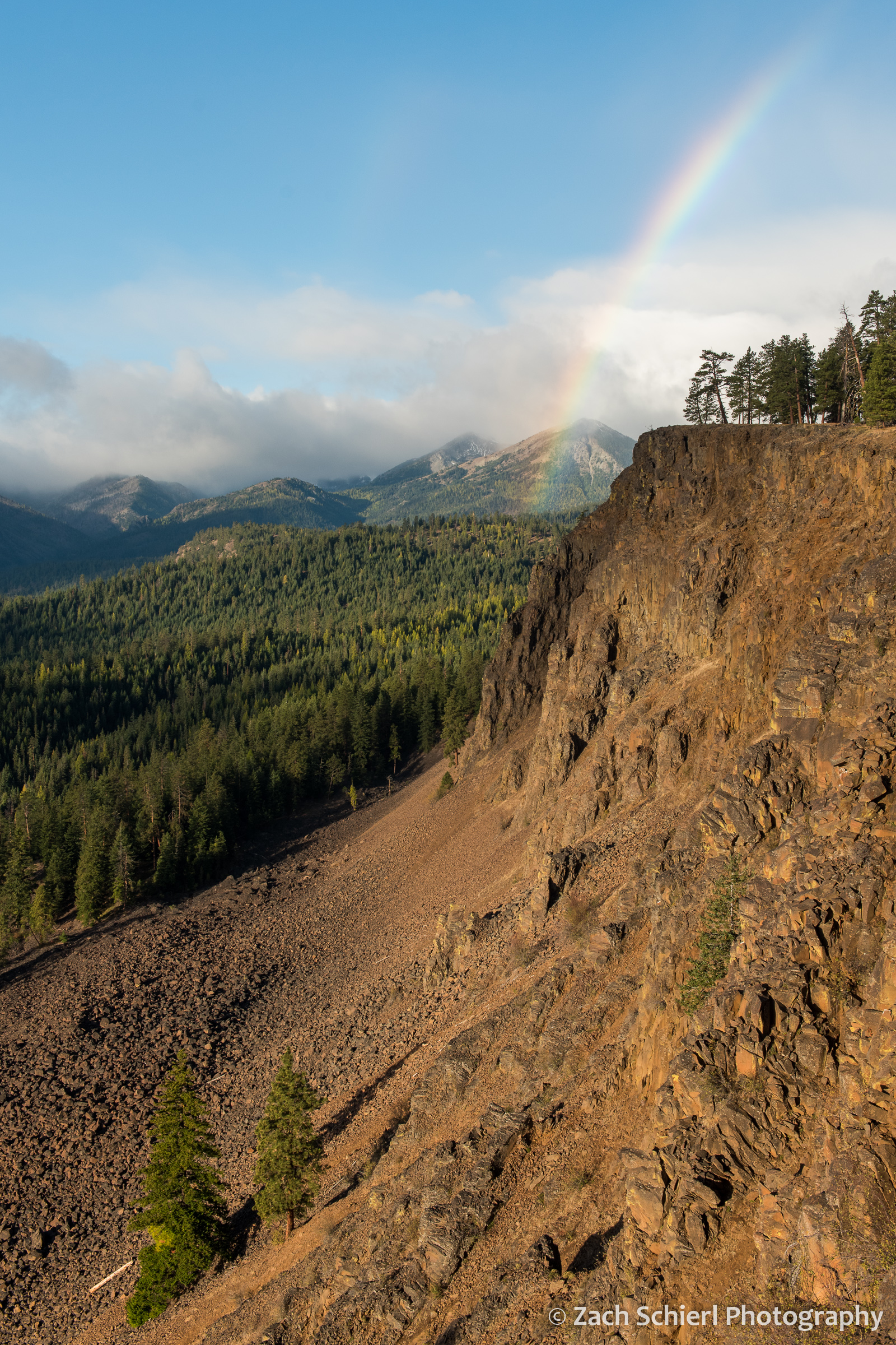 A mountain scene with low clouds, a rainbow, and red-brown cliffs of crumbly rock