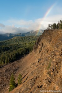 A mountain scene with low clouds, a rainbow, and red-brown cliffs of crumbly rock