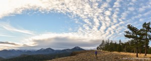 A hiker walks along a grassy meadow as clouds fill the sky overhead