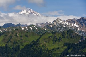 A tall mountain peak is just visible above clouds, with green meadows and forests belows.
