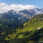A tall mountain peak is just visible above clouds, with green meadows and forests belows.