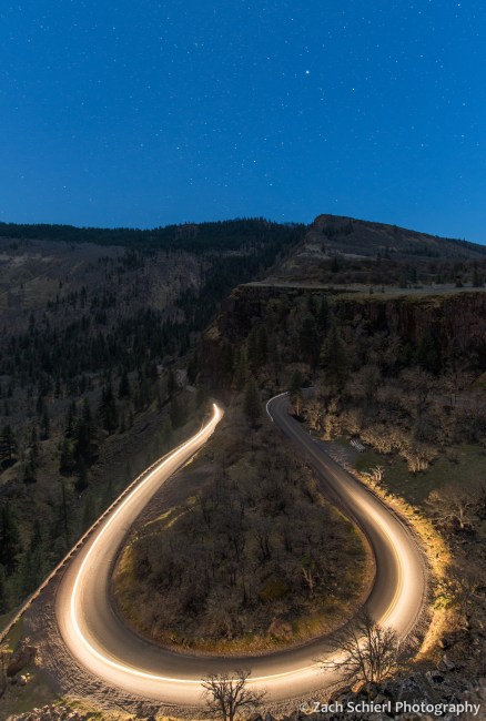 A streak of headlights illuminates a winding mountain road with stars overhead. 