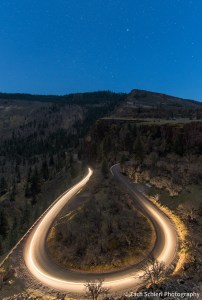 A streak of headlights illuminates a winding mountain road with stars overhead. 