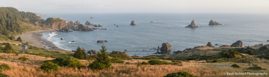 A panoramic view of a grassy slope, a sandy beach, blue ocean, and several rocky islands