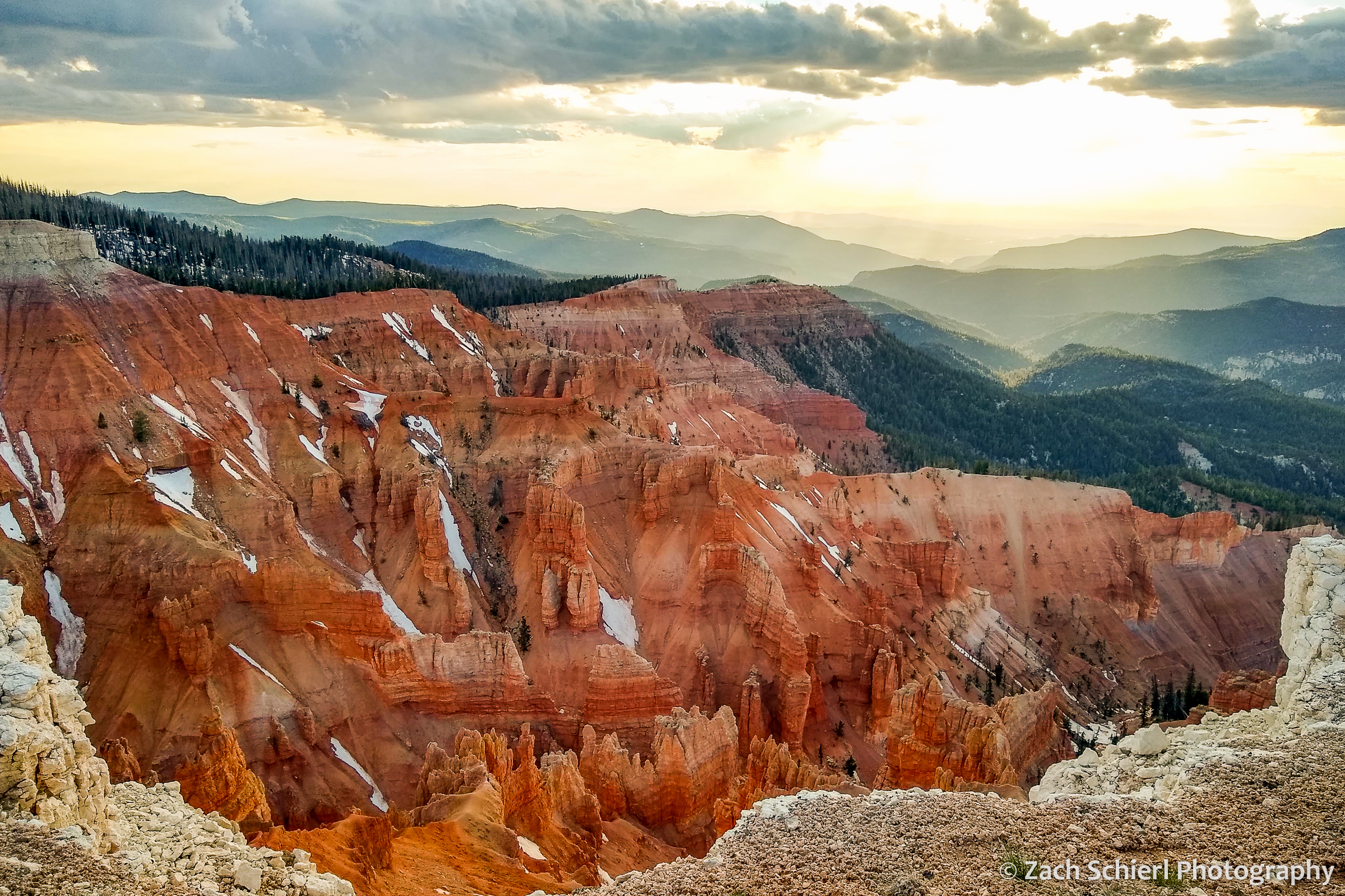 Cliffs of pink, orange, and white rock, with patches of snow and a forest in the background