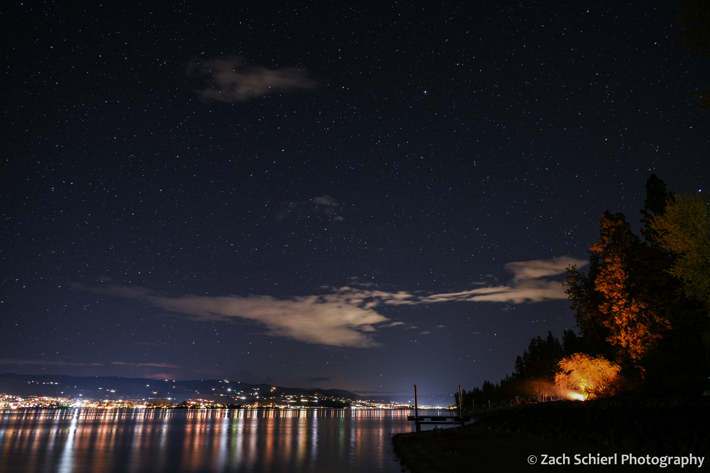 City lights are seen reflected in a lake, while the light of a campfire illuminates trees along the shore.