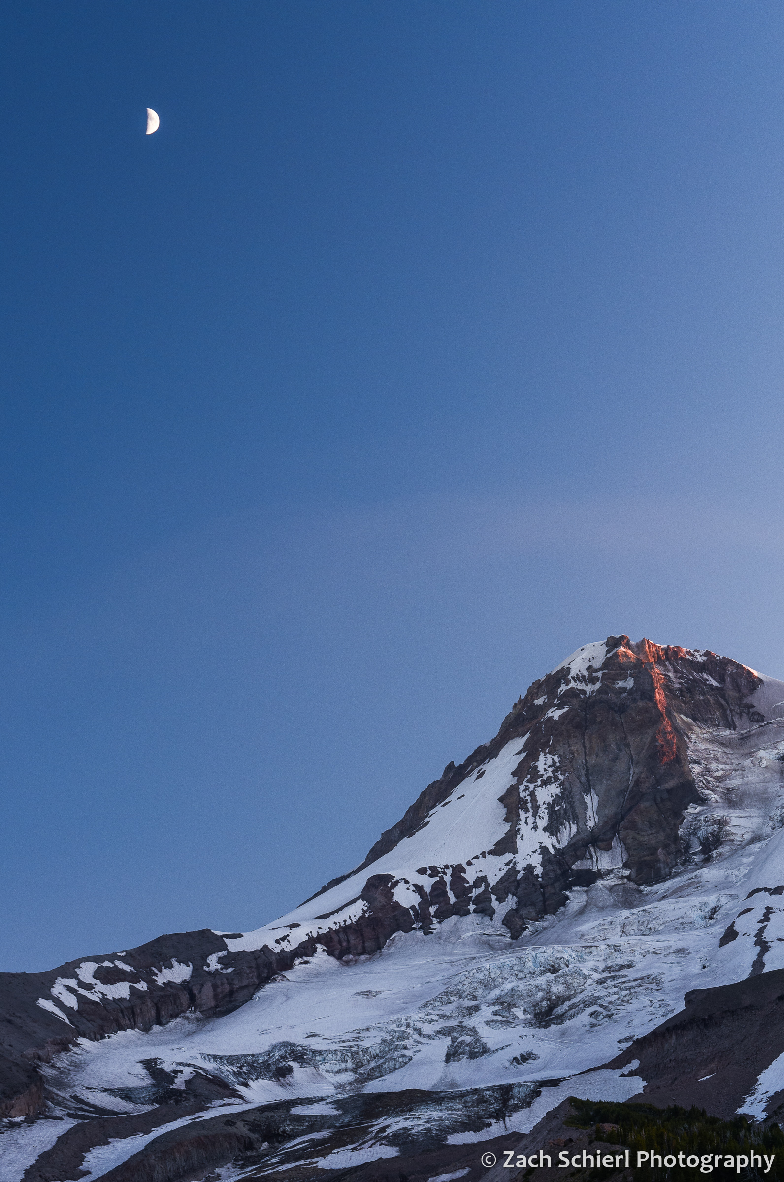 A tall mountain with steep cliffs and ice, with blue sky and a half moon in the sky above it