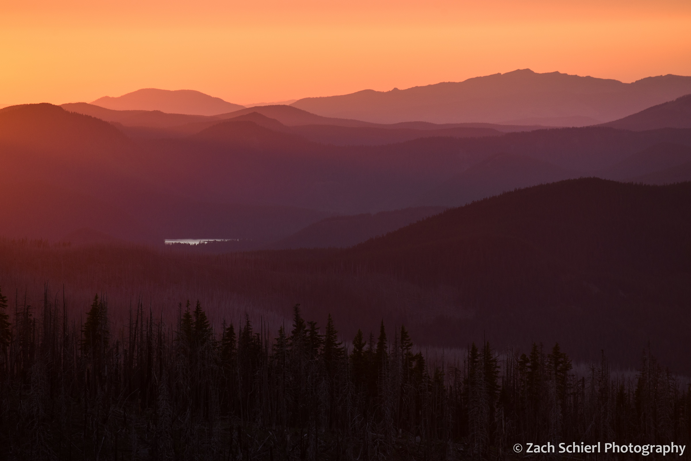 A landscape of forests and hills turns various shades of orange and purple at sunset