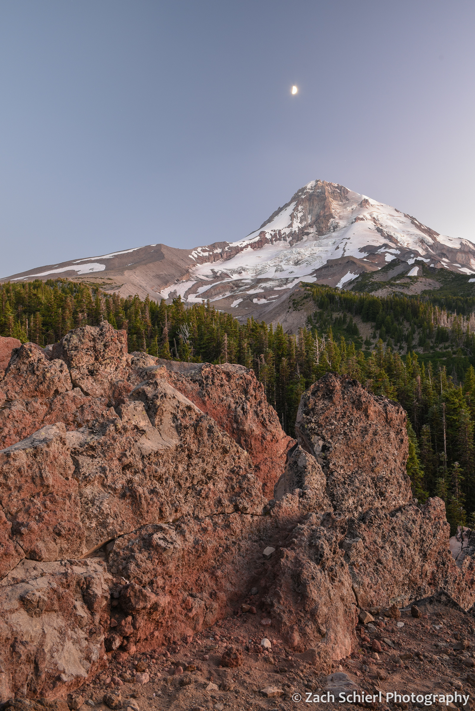 Jagged rocks in the foreground with a tall glacier-capped mountain and quarter moon in the sky