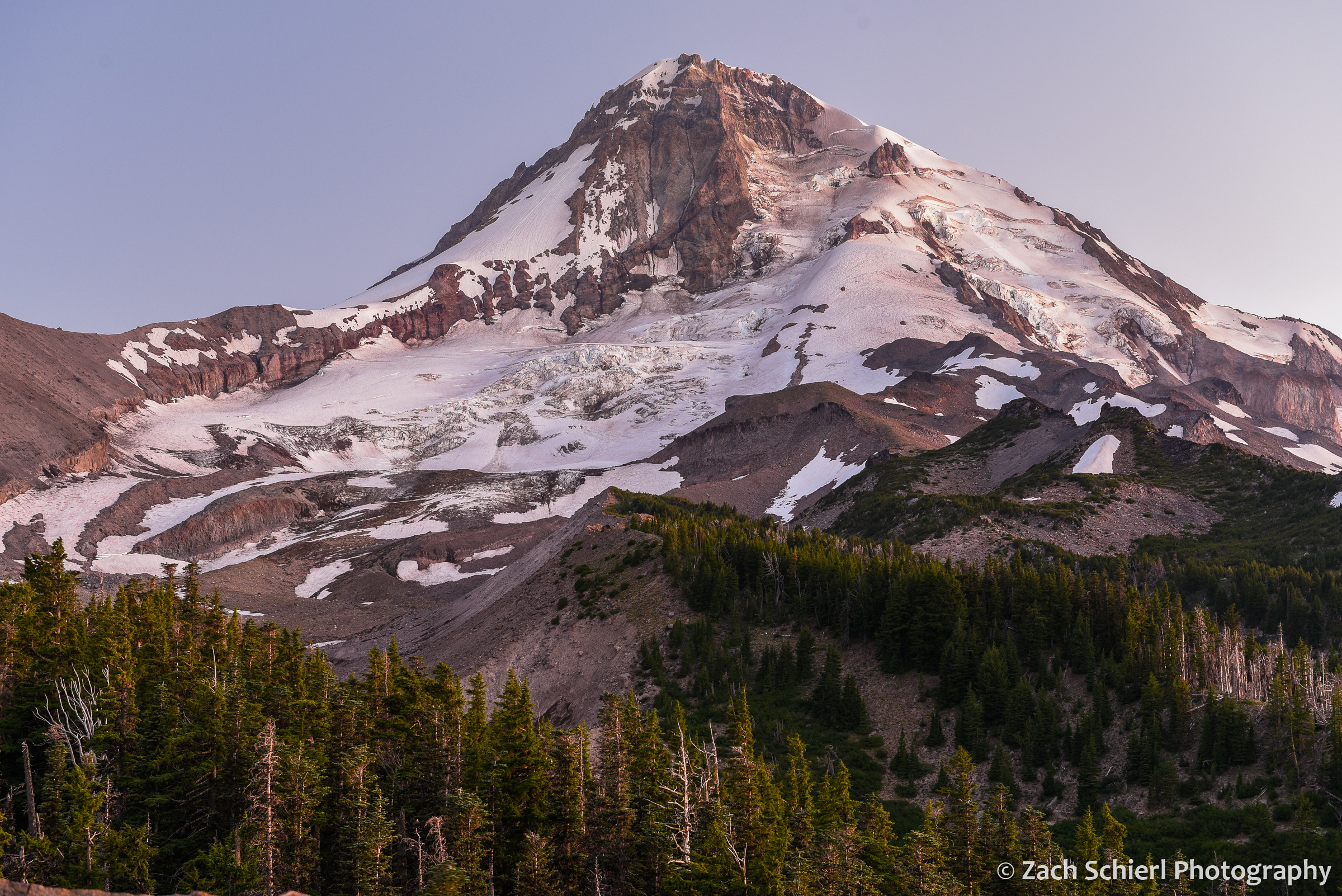 A tall cone-shaped peak with a large white glacier and green trees in the foreground