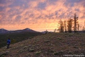 A backpacker looks out an at orange sunset sky while standing on a rocky mesa covered in wildflowers