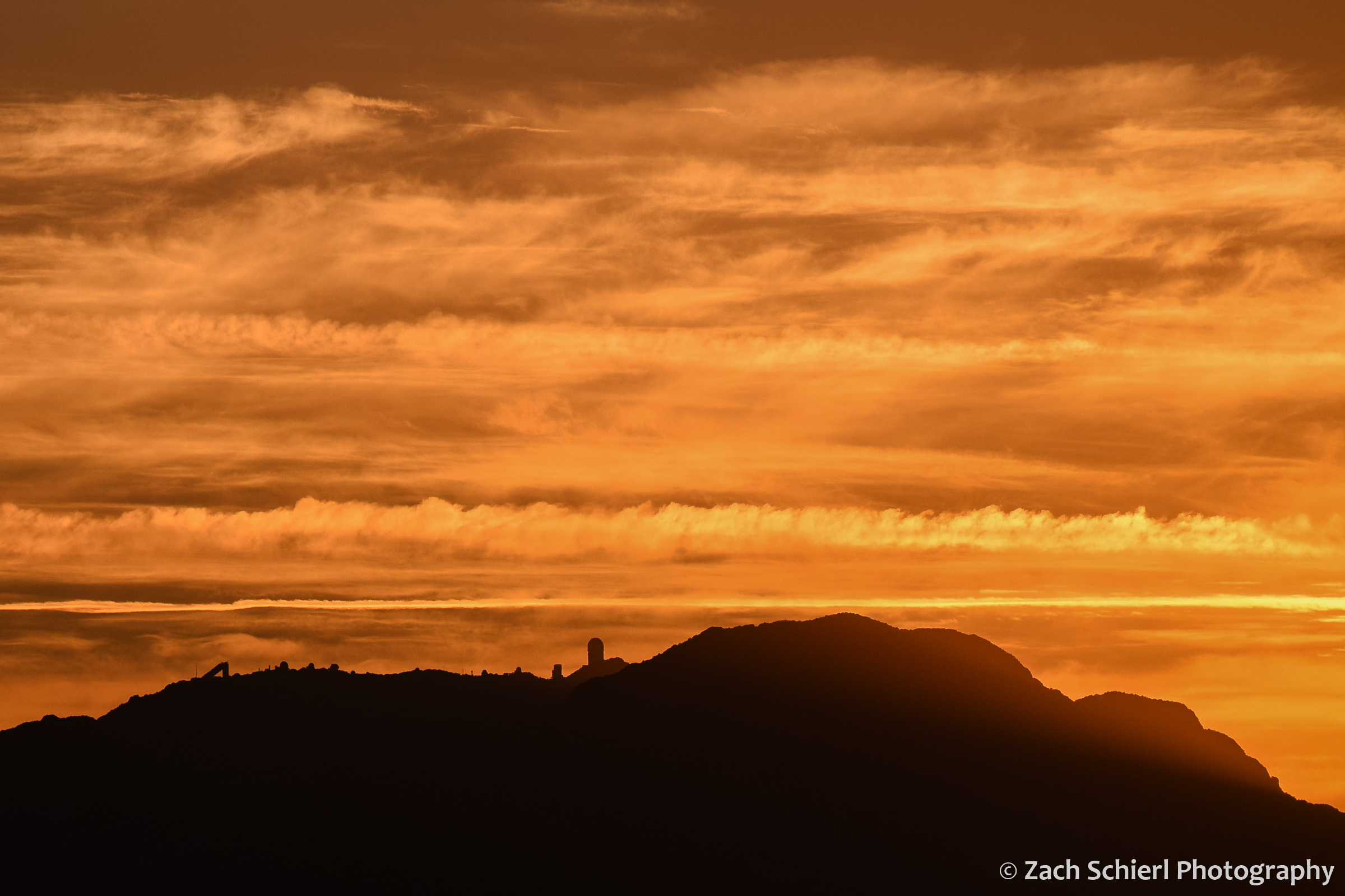 The black silhouette of a mountain is superimposed against a bright orange sky, with several telescope domes along the crest of the mountain